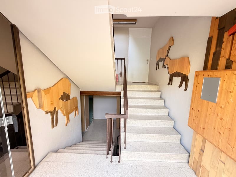 Staircase with white steps and a metal railing, wooden wall-mounted sculptures, and a partially visible room at the top.