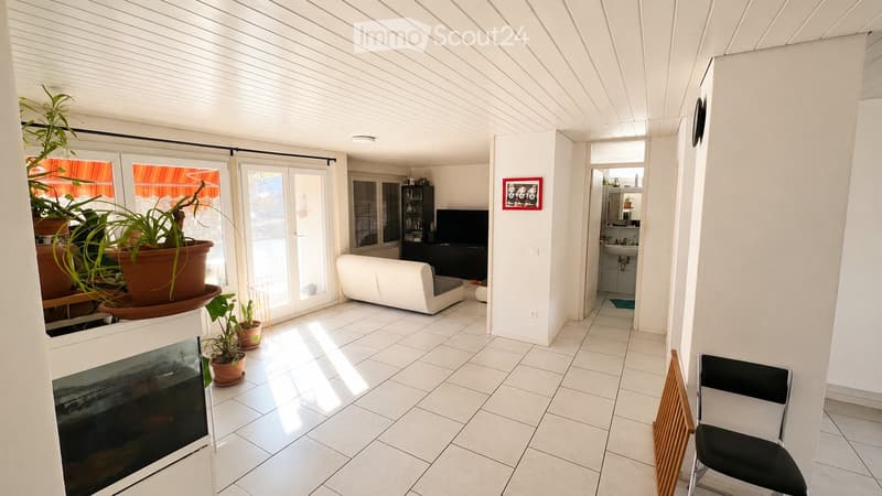 Bright living room with white tiled flooring, white walls, and a white sofa. Features include a large window with blinds, potted plants, a television, and a small table with a chair.