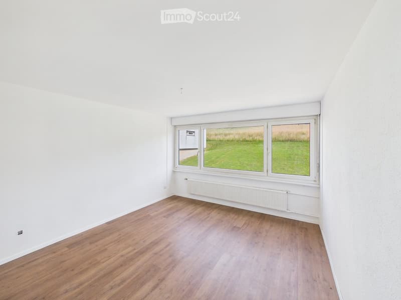 Empty room with white walls, wooden flooring, and large windows overlooking a grassy area.