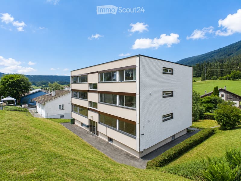 Modern two-story building with white brick facade and large windows, situated on a grassy slope with surrounding greenery and distant hills.