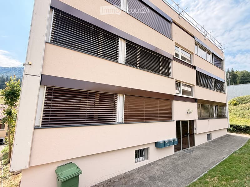 Modern multi-story building with beige facade, large windows with blinds, and a small balcony on the top floor.