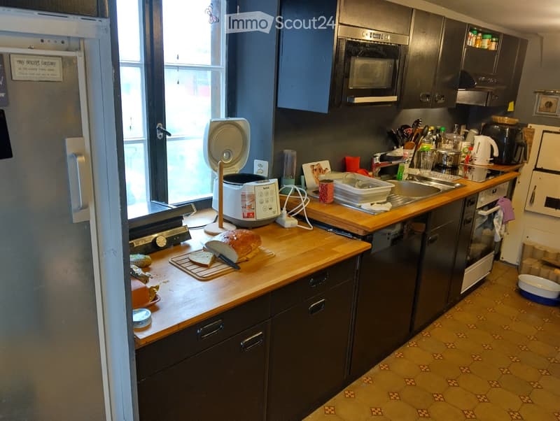 Modern kitchen with dark cabinets, wooden countertops, and various appliances including a microwave, oven, dishwasher, and sink. Bread and a knife are on the counter, along with a fan and other small items.