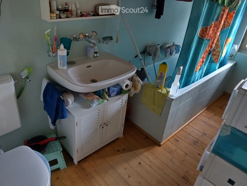 Bathroom with a sink, faucet, toilet, bathtub, and a small cabinet. The sink has a toothbrush holder and toothpaste, and there are towels and a bottle on the edge of the bathtub. A small stool is visible in the foreground.