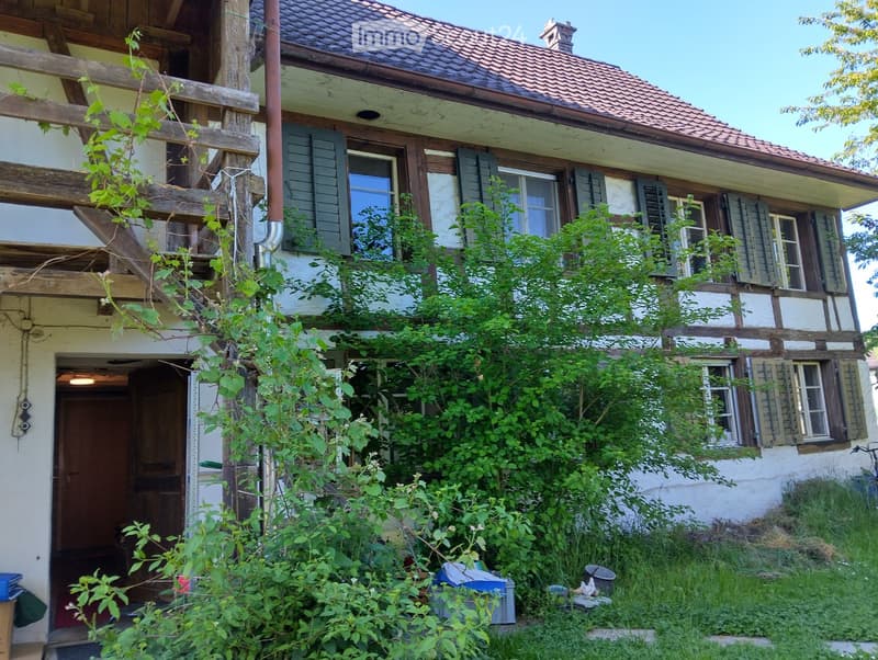 Two-story traditional house with wooden beams, green shutters, and a tiled roof, partially overgrown with vegetation.