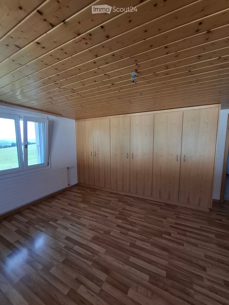 Empty attic room with wooden slatted ceiling, light wooden floor, large window with view, and built-in wooden closet units.