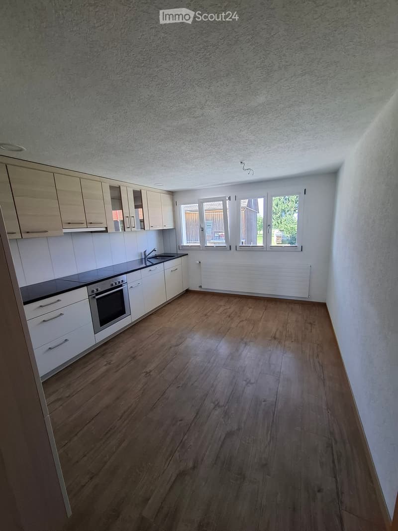 Empty kitchen with light wooden cabinets, white tiled backsplash, and wooden flooring. Windows provide natural light, and the space is unfurnished.