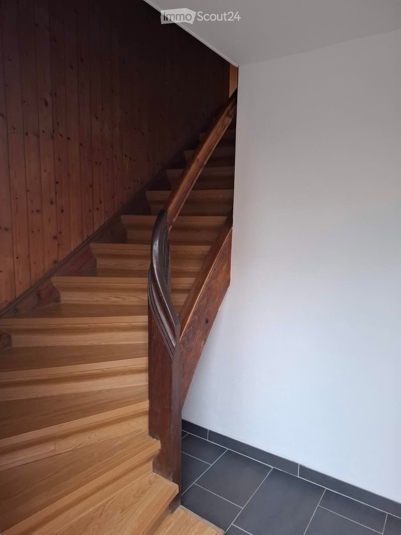 Wooden staircase with a dark wooden handrail and light wooden steps, leading up to a higher level. The walls are painted white, and the lower part of the wall is tiled in dark gray tiles.