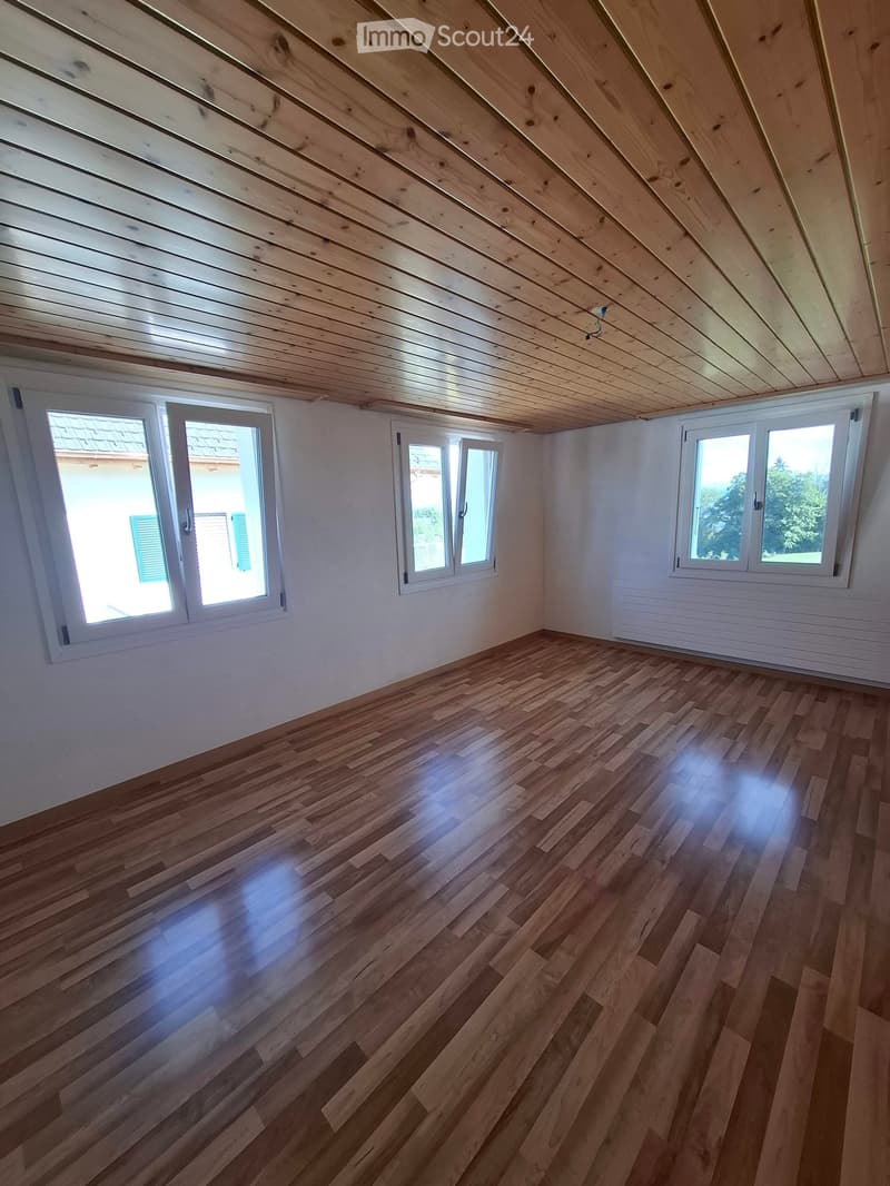 Empty attic room with wooden floor and slanted wooden ceiling, featuring multiple windows with white frames and shutters, natural light flooding the space.