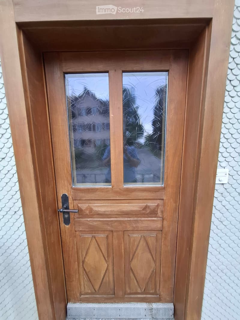 Wooden front door with glass panels and decorative diamond-shaped panels, set in a white textured wall with a small step leading up to it.