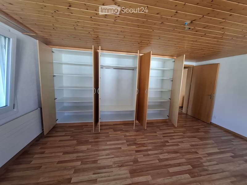 Empty room with wooden flooring, white walls, and built-in wooden closet with multiple shelves and a hanging rod. The room has a large window on one side and a door leading to another room.
