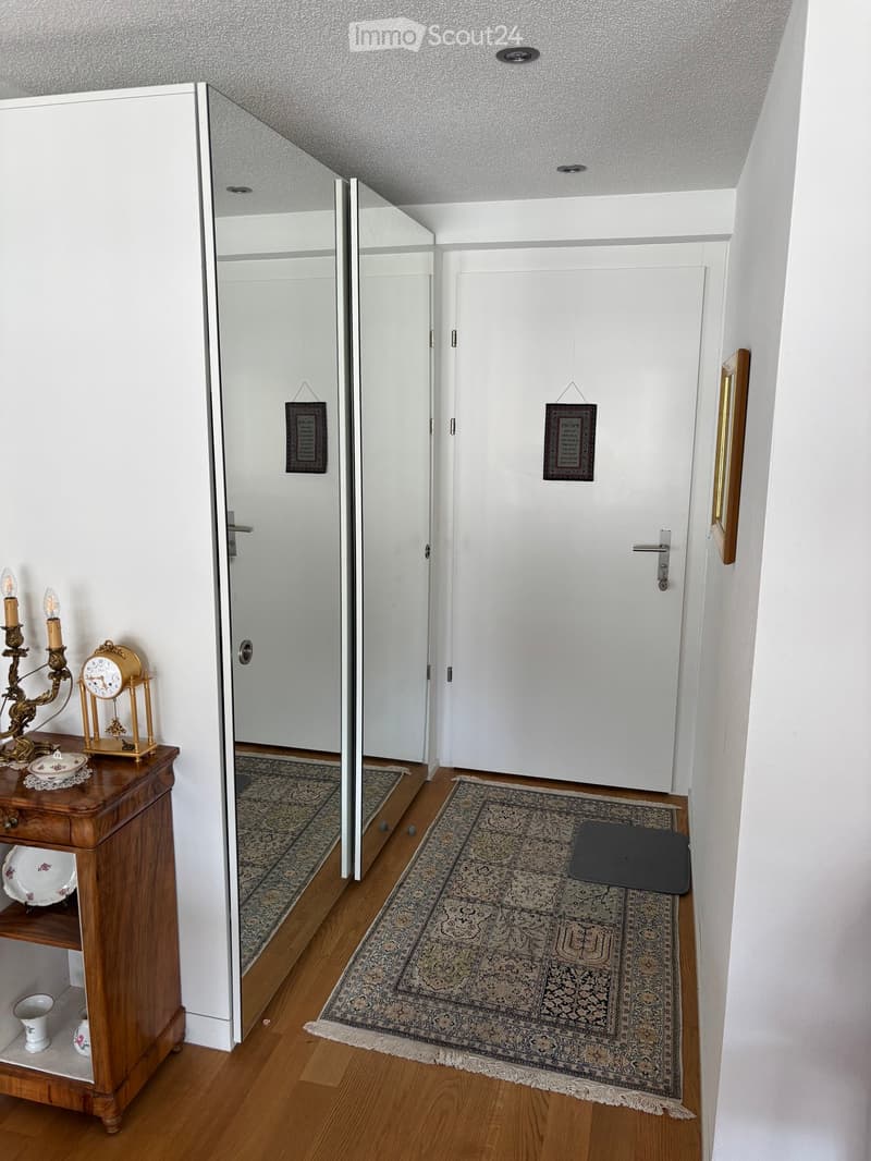 A narrow hallway with wooden flooring, white walls, and two doors. A small wooden table with a clock, candle holder, and decorative items is placed on the left side. Two rugs are placed in front of the doors.