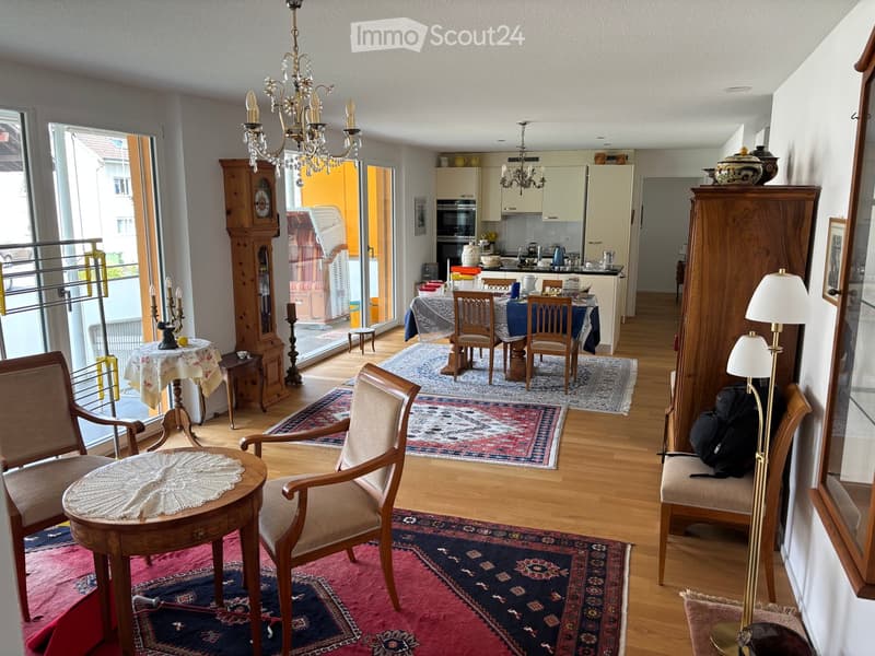 A well-furnished living room with wooden floors, multiple seating areas, a dining area, and a kitchen. Features include a chandelier, a grandfather clock, and various decorative items.