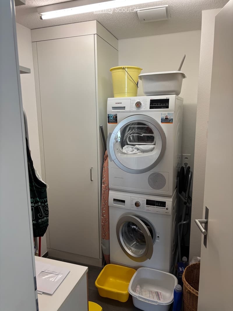 Compact bathroom with a stacked washing machine and dryer, white cabinets, and various storage items including buckets and a wicker basket.