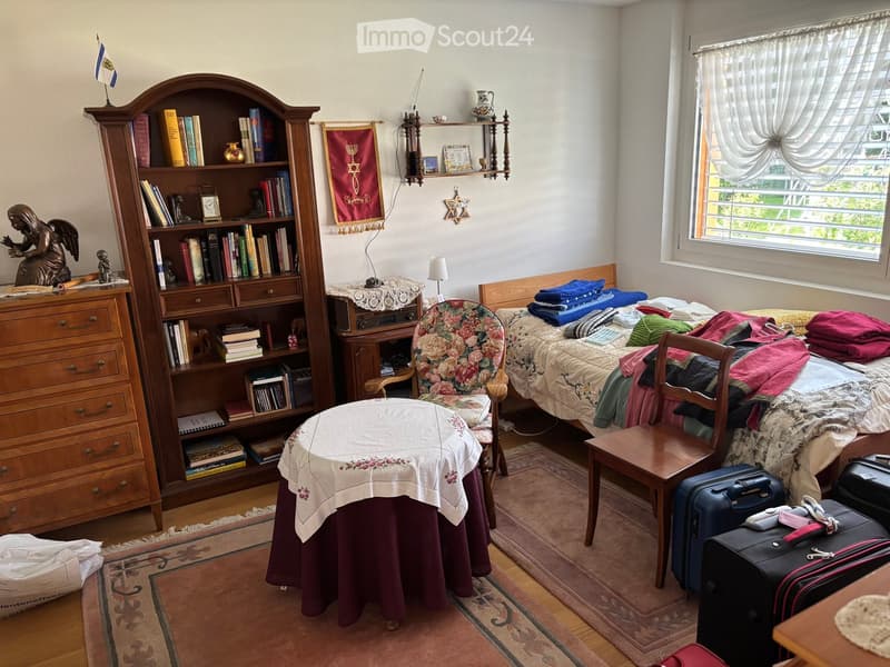 A cozy bedroom with a double bed, a wooden bookshelf filled with books, a small table with a floral tablecloth, a wooden chair, and a suitcase on the floor. The room features a window with white curtains and a decorative wall hanging.