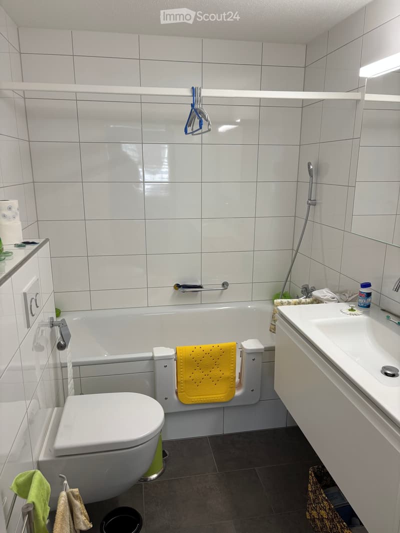 Modern bathroom with white tiles, bathtub, toilet, sink, and mirror. Includes a yellow towel on the edge of the bathtub and a green towel on the floor.