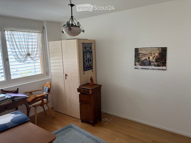 A furnished room with wooden flooring, a window with curtains, a wooden chair with a cushion, a small table with books, a wooden cabinet, a decorative picture frame, and a small rug.