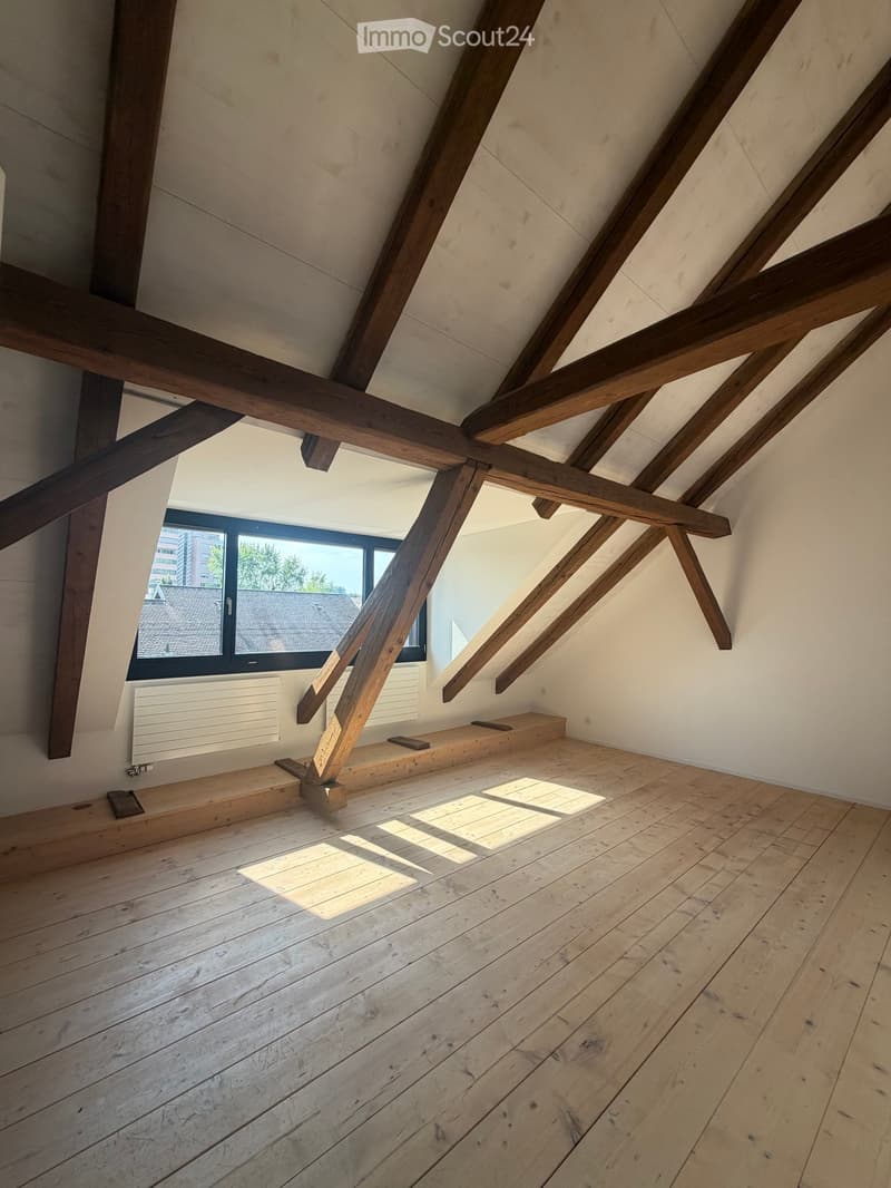 An empty attic space with wooden beams, wooden flooring, and a large window offering a view of the outside. The room features exposed wooden beams and a radiator on one wall.