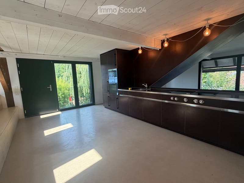 Modern kitchen with dark wooden cabinets, stainless steel appliances, and a large island. The space features a concrete floor and exposed wooden ceiling beams.