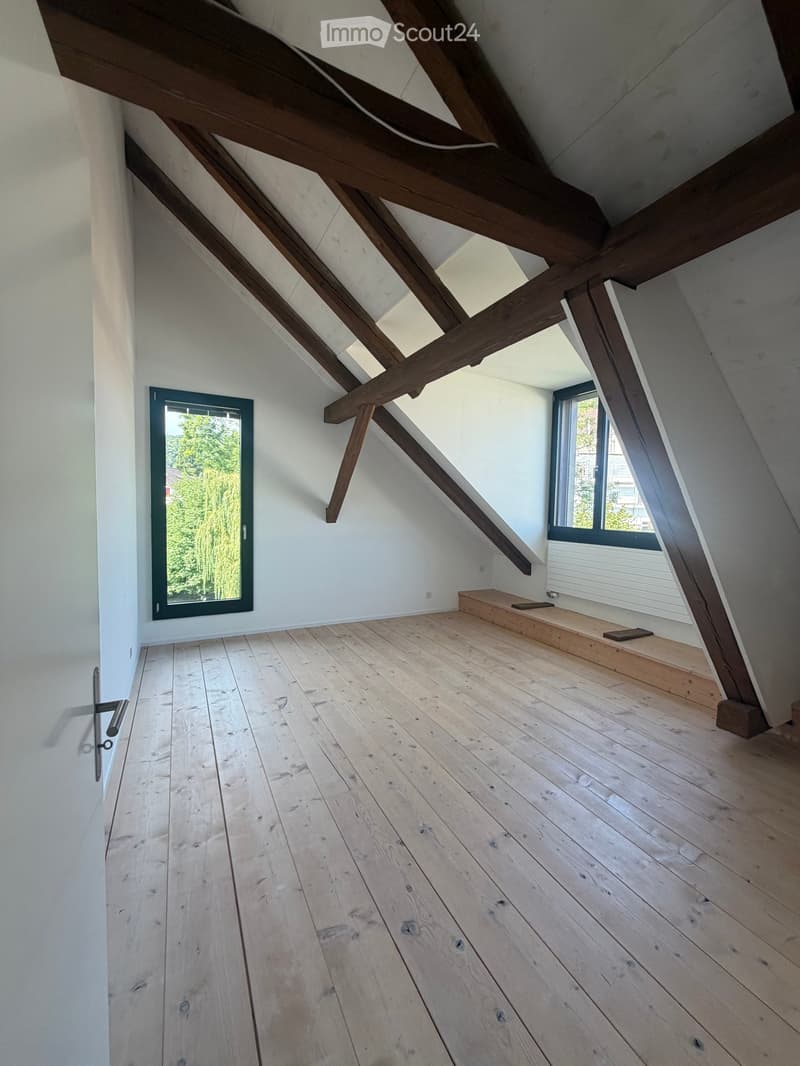 An empty attic space with wooden beams, light wooden flooring, and two windows offering a view of greenery outside.