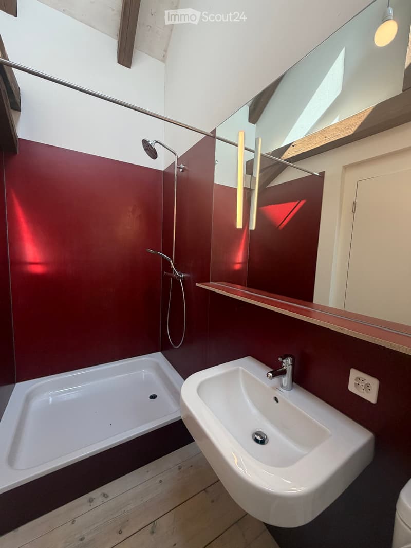 Modern bathroom with a white bathtub, white sink, chrome faucet, and a large mirror above the sink. The walls are painted in a deep red color, and the floor is made of light wood.