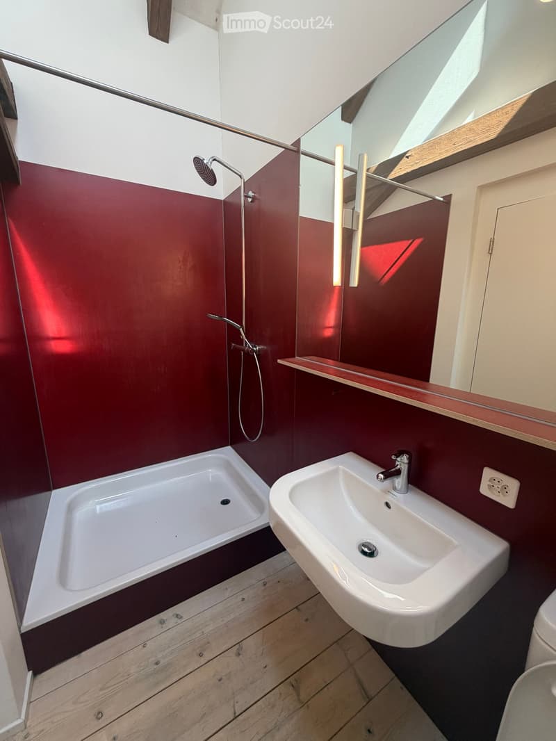 Modern bathroom with red walls, white bathtub, white sink, and a showerhead on the wall. Features a large mirror and wooden flooring.