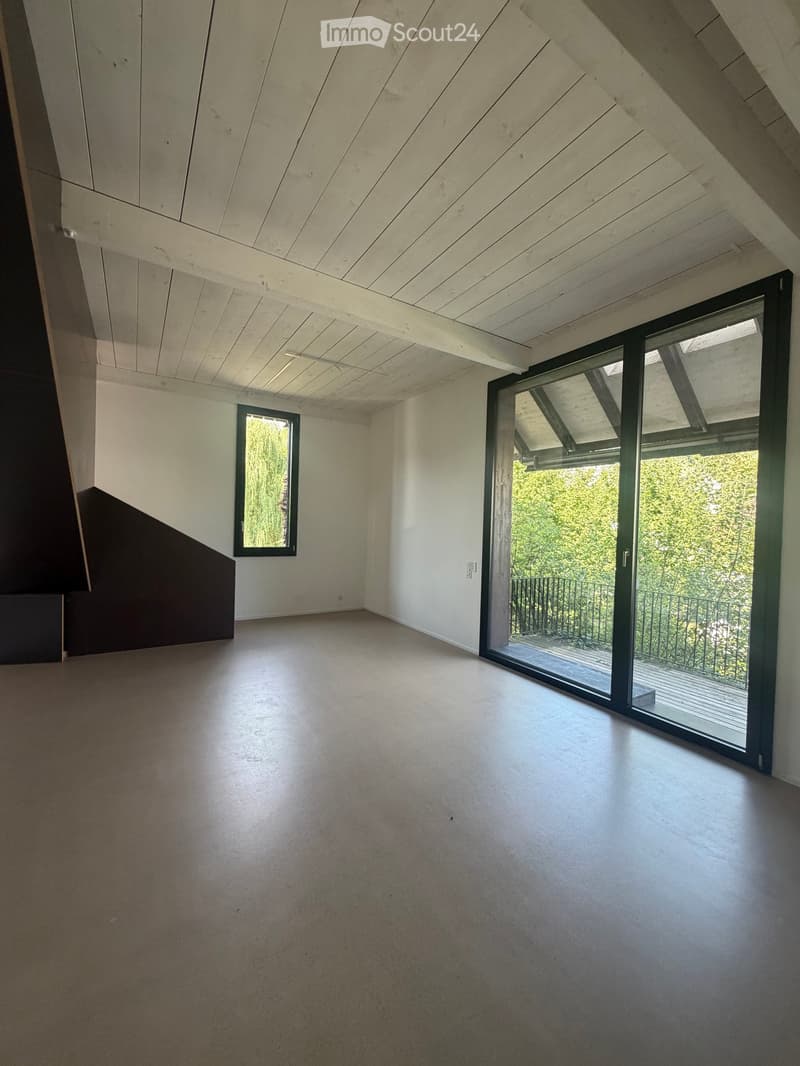 An empty room with a wooden slatted ceiling, large sliding glass door leading to a balcony, and a small window on the left side.
