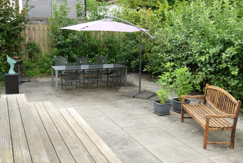 A well-maintained garden patio with a wooden bench, a dining table with chairs, potted plants, and a large umbrella providing shade.