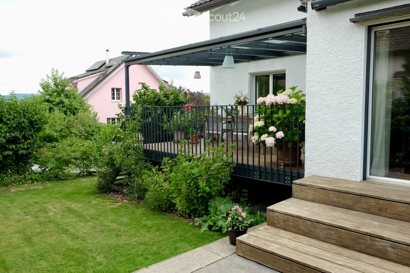 Garden with a well-maintained lawn, wooden steps leading to a covered balcony area, potted plants, and a modern house with large windows.