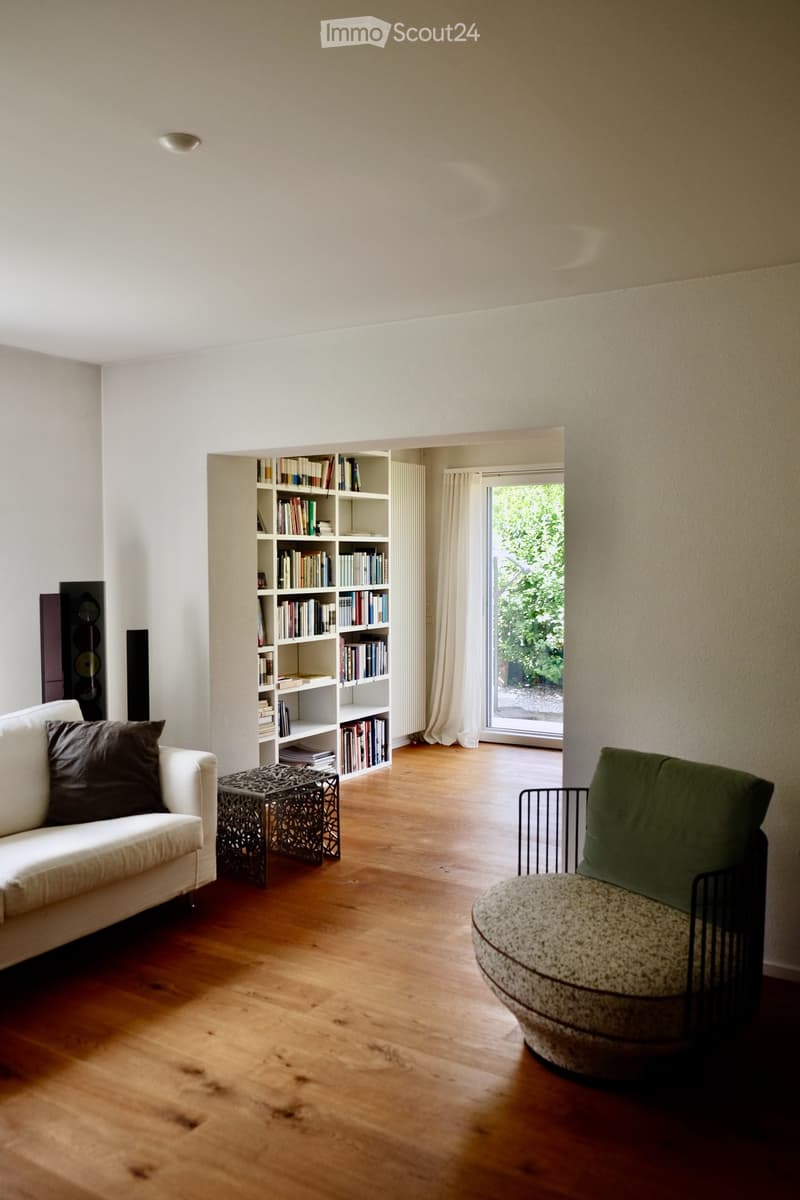 Modern living room with light wooden flooring, white walls, a white sofa with brown cushions, a small decorative table, a round chair, a bookshelf filled with books, and a large window with white curtains leading to a garden view.