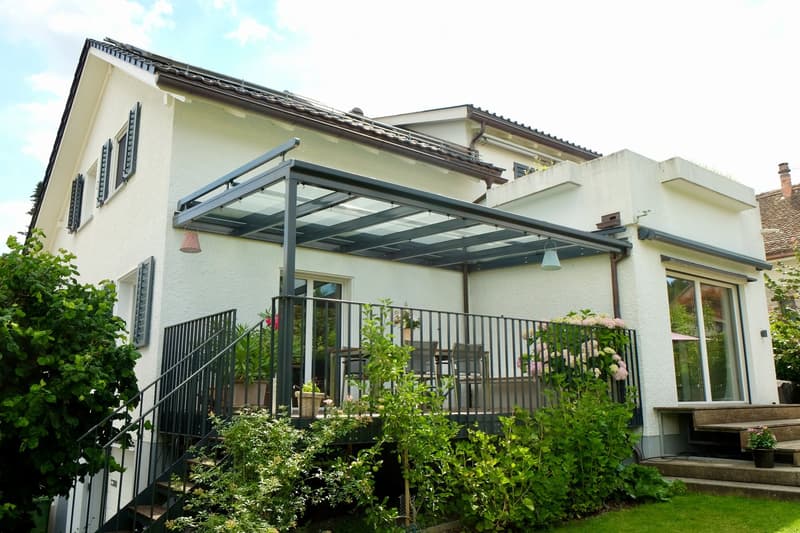 Two-story house with a covered patio and balcony, surrounded by greenery and potted plants.