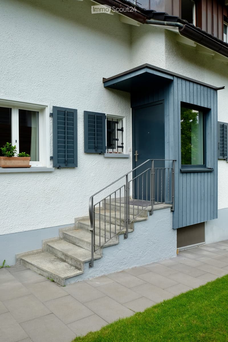 Two-story house with white textured walls, grey shutters, and a small balcony with a potted plant. A staircase with a metal railing leads to the entrance door.