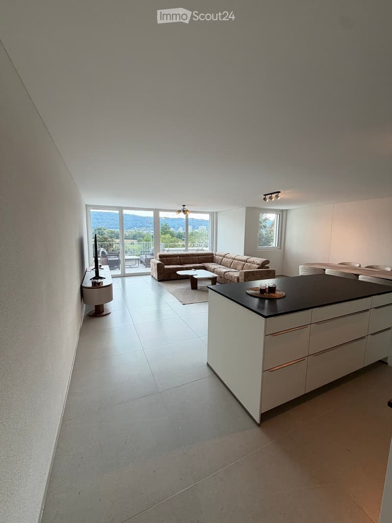 Modern open-plan living and dining area with a sectional sofa, coffee table, kitchen island, and large windows offering a scenic view.