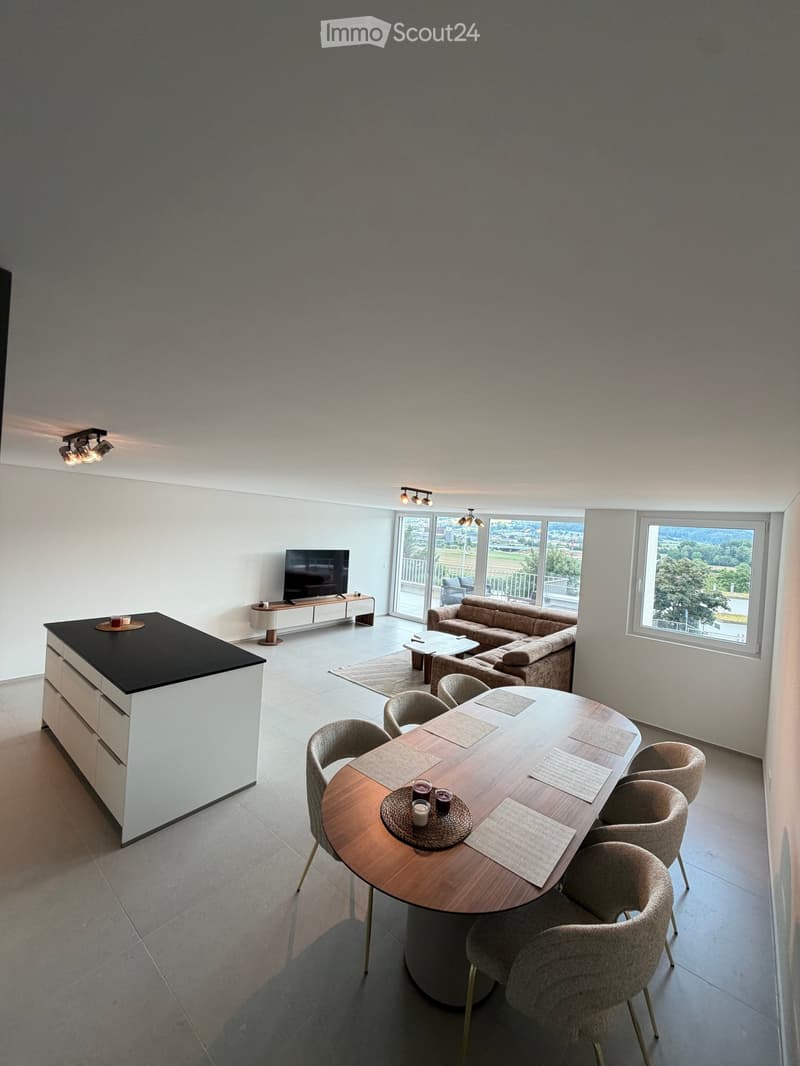 Modern living and dining area with a sofa, coffee table, dining table with chairs, and a kitchen island. Large windows offer a scenic view.