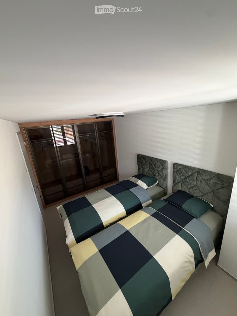 A tidy bedroom with two single beds, each featuring a grey headboard and matching bedding in a geometric pattern of green, blue, and cream. A large mirrored wardrobe with wooden framing is positioned against the wall.