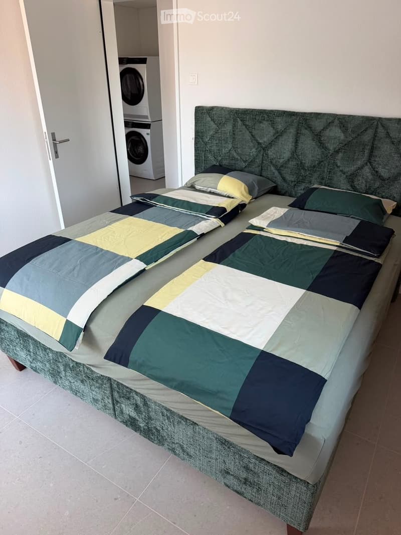 A modern bedroom with a large bed featuring a geometric duvet in shades of green, blue, and yellow, complemented by matching pillows. A washing machine is visible in an adjacent closet area.