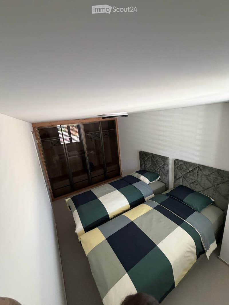A modern bedroom with a large bed featuring a geometric duvet in green, blue, and beige, flanked by matching pillows. A built-in wardrobe with mirrored doors is visible on the left side.
