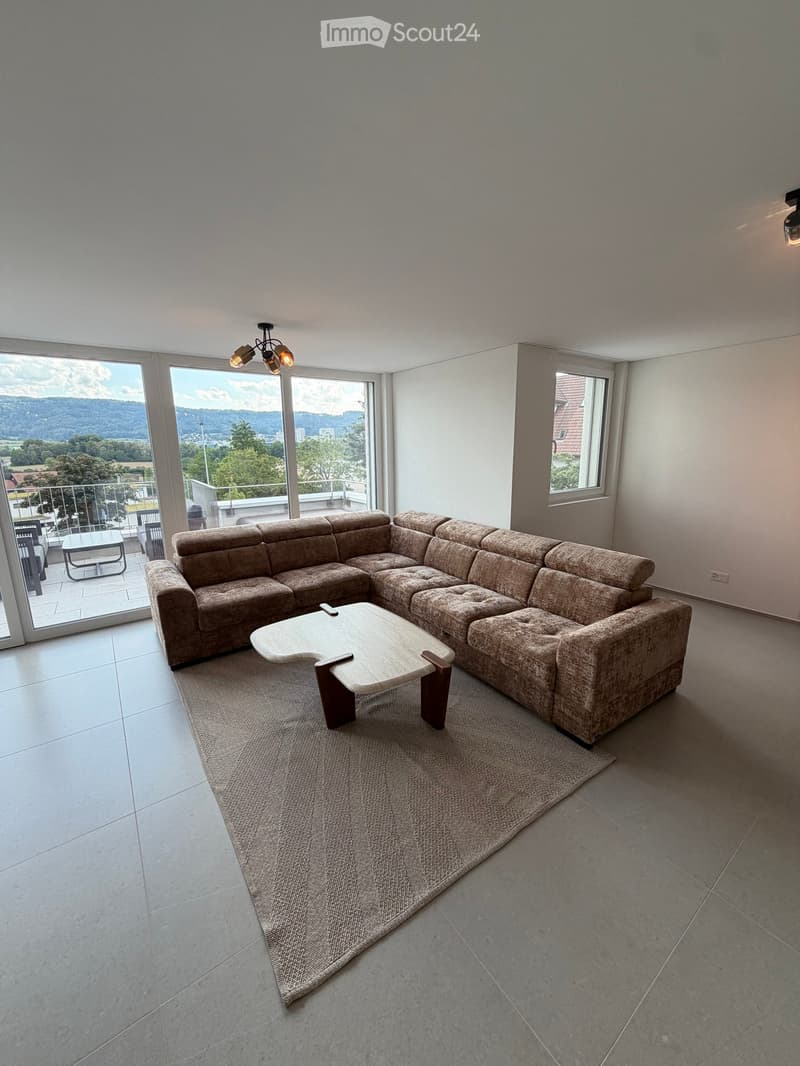 Modern living room with large windows offering a scenic view, featuring a sectional sofa, coffee table, and a small table with chairs on a balcony.