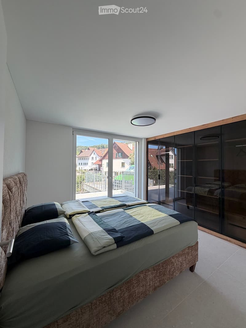 Modern bedroom with a large bed, patterned bedding, and a sliding glass door leading to a balcony with a view of the neighborhood.