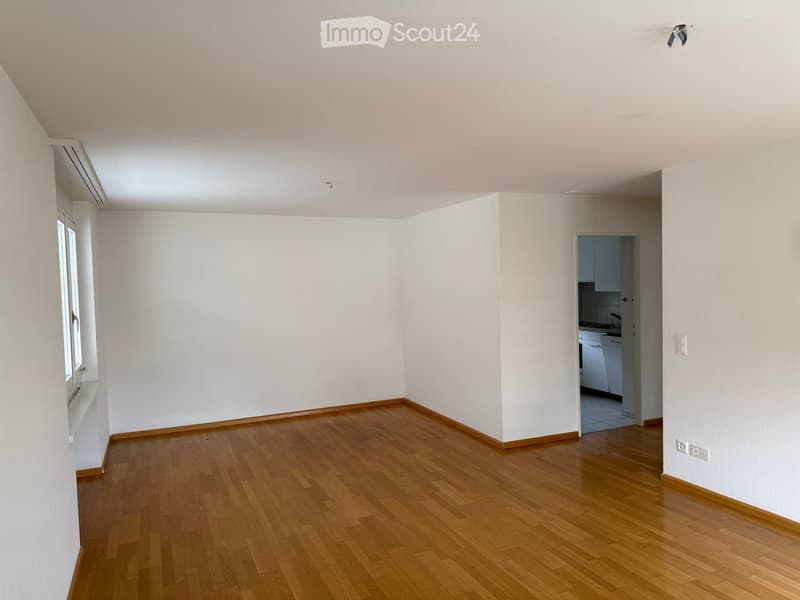 Empty room with wooden flooring, white walls, and a window on one side. A small kitchen area is visible through an open doorway.