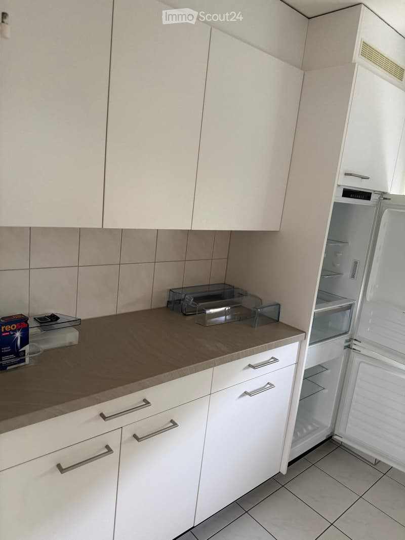 Modern kitchen with white cabinets, a countertop, and an open refrigerator. The countertop has a few items, including a box and a smartphone.
