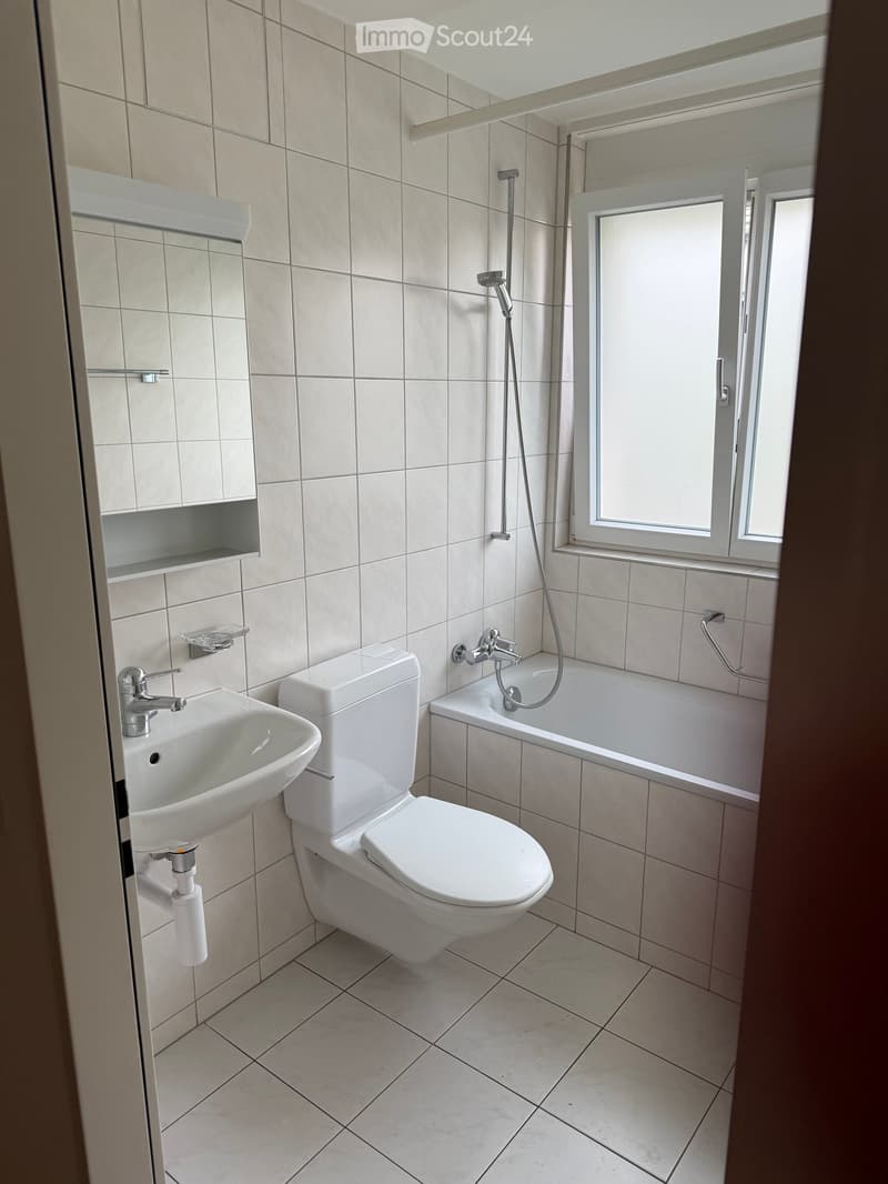 Modern bathroom with white tiles, a sink, toilet, bathtub, and a window with blinds.