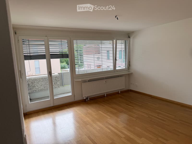 Empty room with wooden flooring, large windows with blinds, radiator, and a sliding glass door leading to a balcony area.