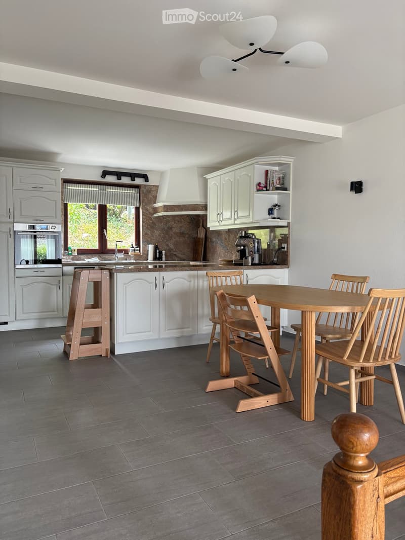 Modern kitchen with white cabinets, a central dining table with chairs, and a built-in oven and fridge. Features a coffee machine on the counter and a window with blinds.