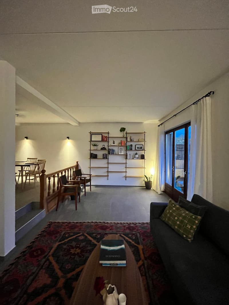 A modern living room with a dark grey sofa, coffee table, books, shelves, and a dining area visible in the background.