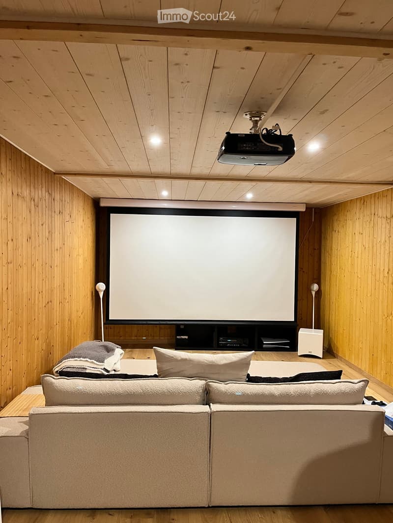 Modern home theater room with wooden walls and ceiling, large screen, projector, sofa, pillows, and audio system.