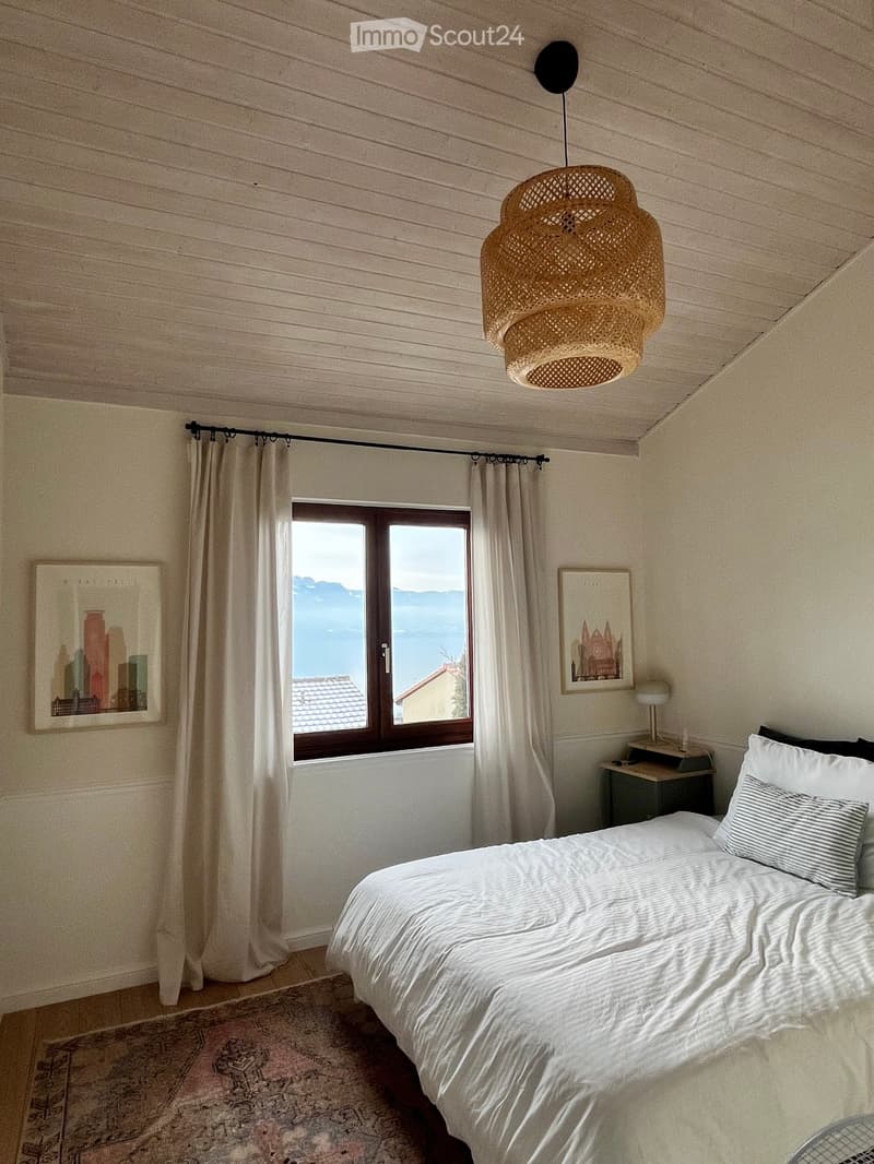 Cozy bedroom with a large bed, nightstand, table lamp, and scenic view through a window. Features a wicker pendant light and decorative picture frames.