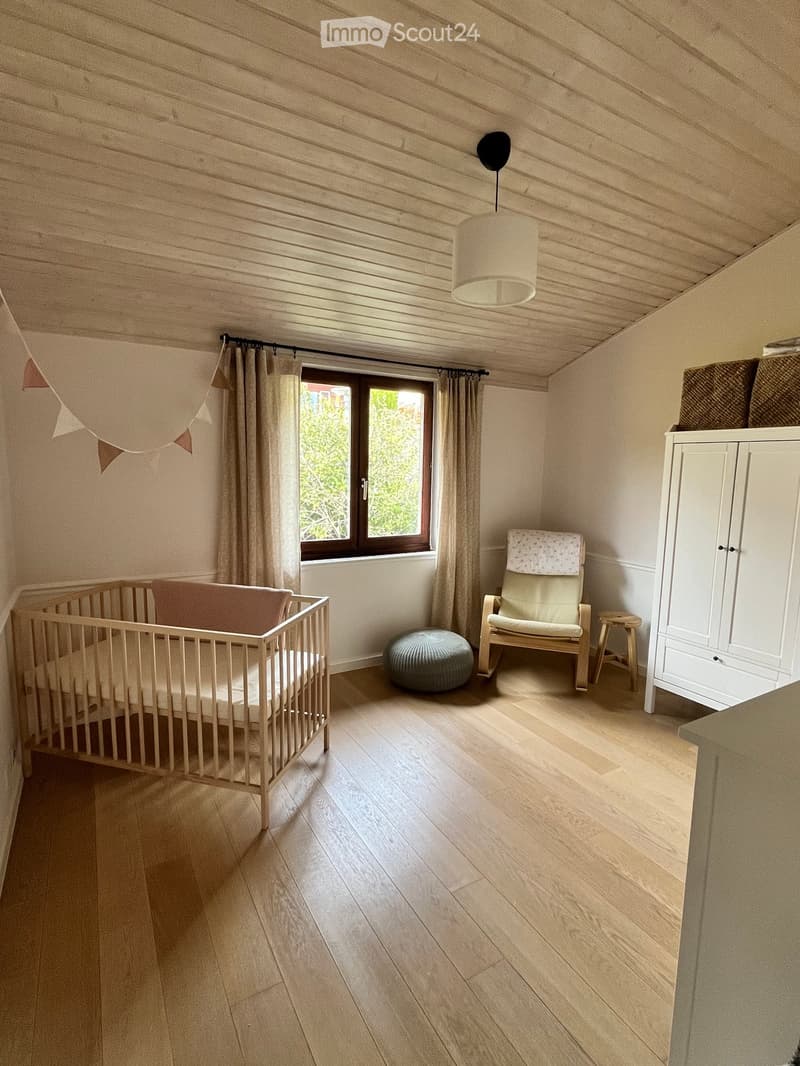 Bright, cozy bedroom with wooden flooring, a crib, a white cabinet, a hanging pendant light, a small chair, and a round ottoman.