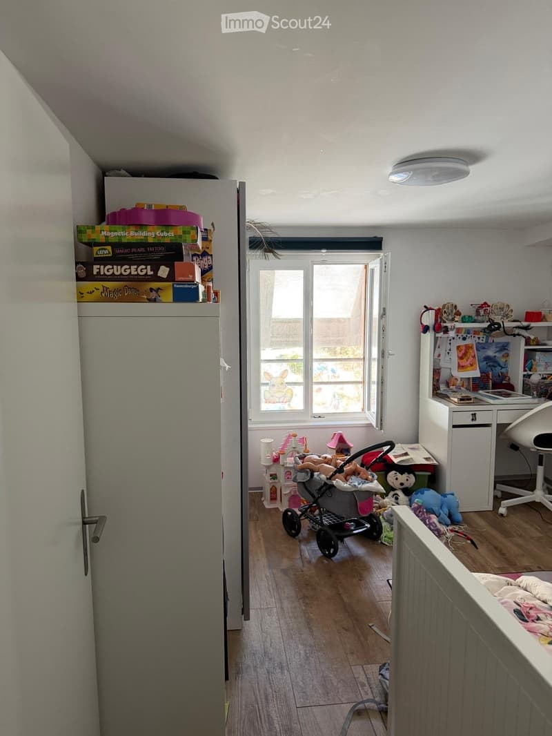 A well-lit room with wooden flooring, a baby stroller, a desk with a chair, and various toys scattered around. A refrigerator is visible on the left, and a window with blinds is open to let in natural light.