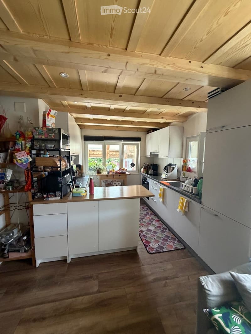 The image shows a modern kitchen with white cabinets, a wooden countertop, and a central island. The kitchen features a sink, stove, refrigerator, and various appliances. There is a small seating area with a table and chairs, and a view of a garden throug