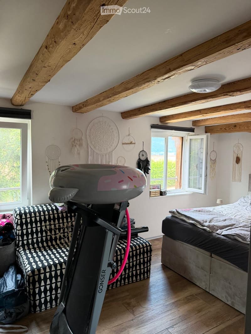 Cozy bedroom with exposed wooden beams, a large bed, a patterned sofa, an exercise bike, and dreamcatchers on the walls. Large windows provide natural light and a view of the outdoors.